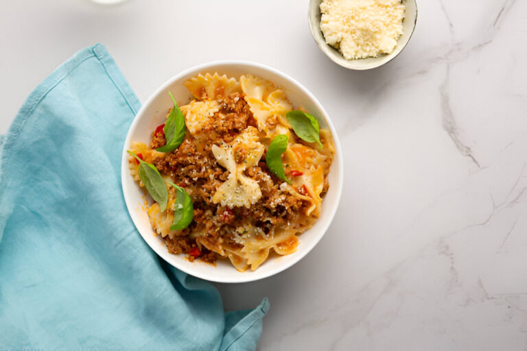 A bowl of pasta with meat sauce and fresh basil leaves, accompanied by a small bowl of ricotta cheese, on a marble countertop with a blue cloth.