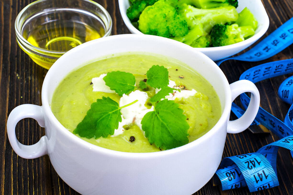 A creamy green soup garnished with fresh herbs in a white bowl, accompanied by steamed broccoli and a bowl of olive oil, with a measuring tape in the foreground.