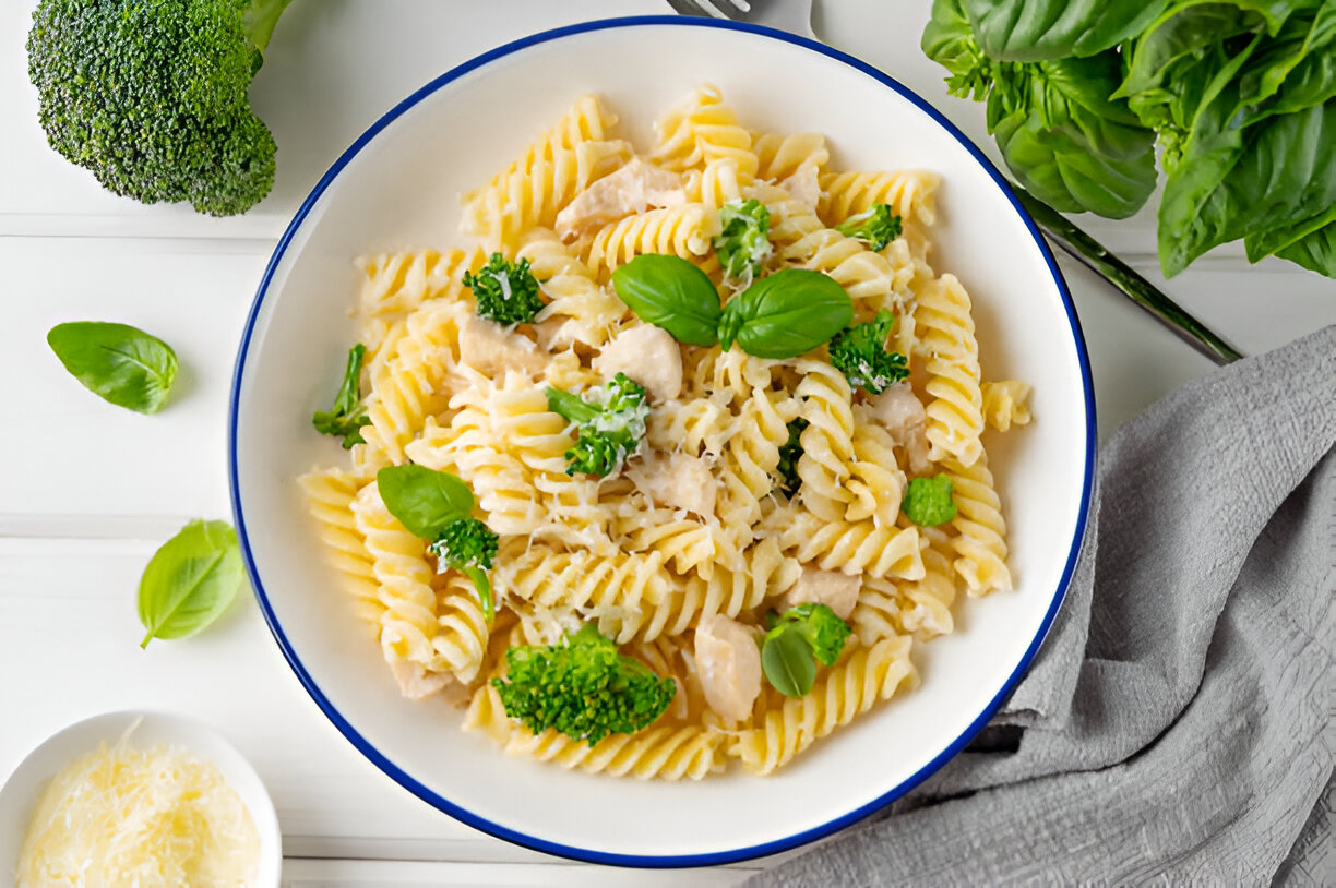 A bowl of creamy pasta with rotini, broccoli, and basil leaves, garnished with shredded cheese.