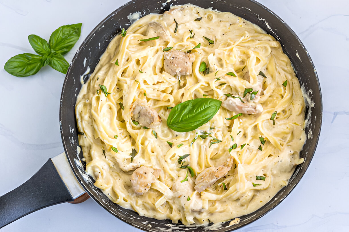 Creamy fettuccine pasta with chicken pieces and fresh basil leaves in a skillet.