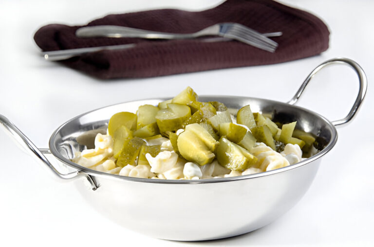 A bowl of pasta topped with chopped pickles, served in a stainless steel dish.