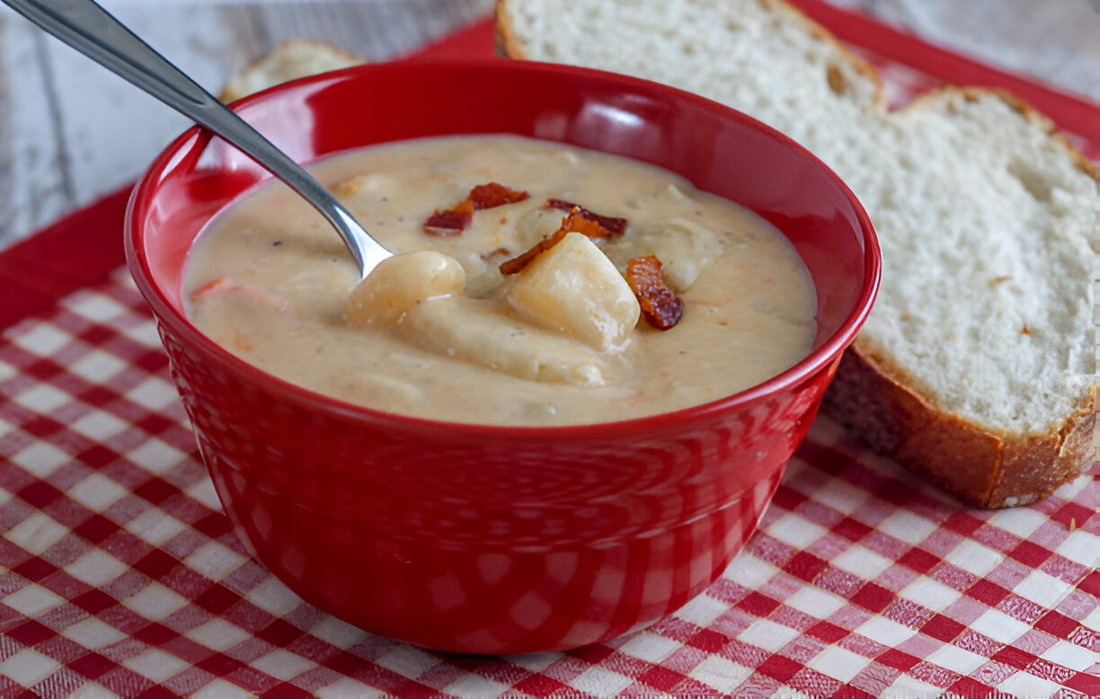 A bowl of creamy soup with bacon bits, served alongside slices of bread on a red checkered tablecloth.