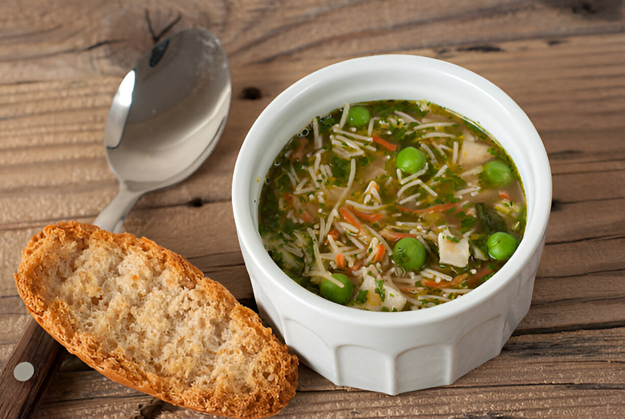 A bowl of vegetable and chicken soup with green peas and a slice of bread on a wooden surface.