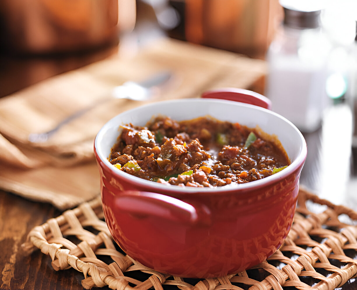 A red bowl filled with chili on a woven mat with utensils in the background.