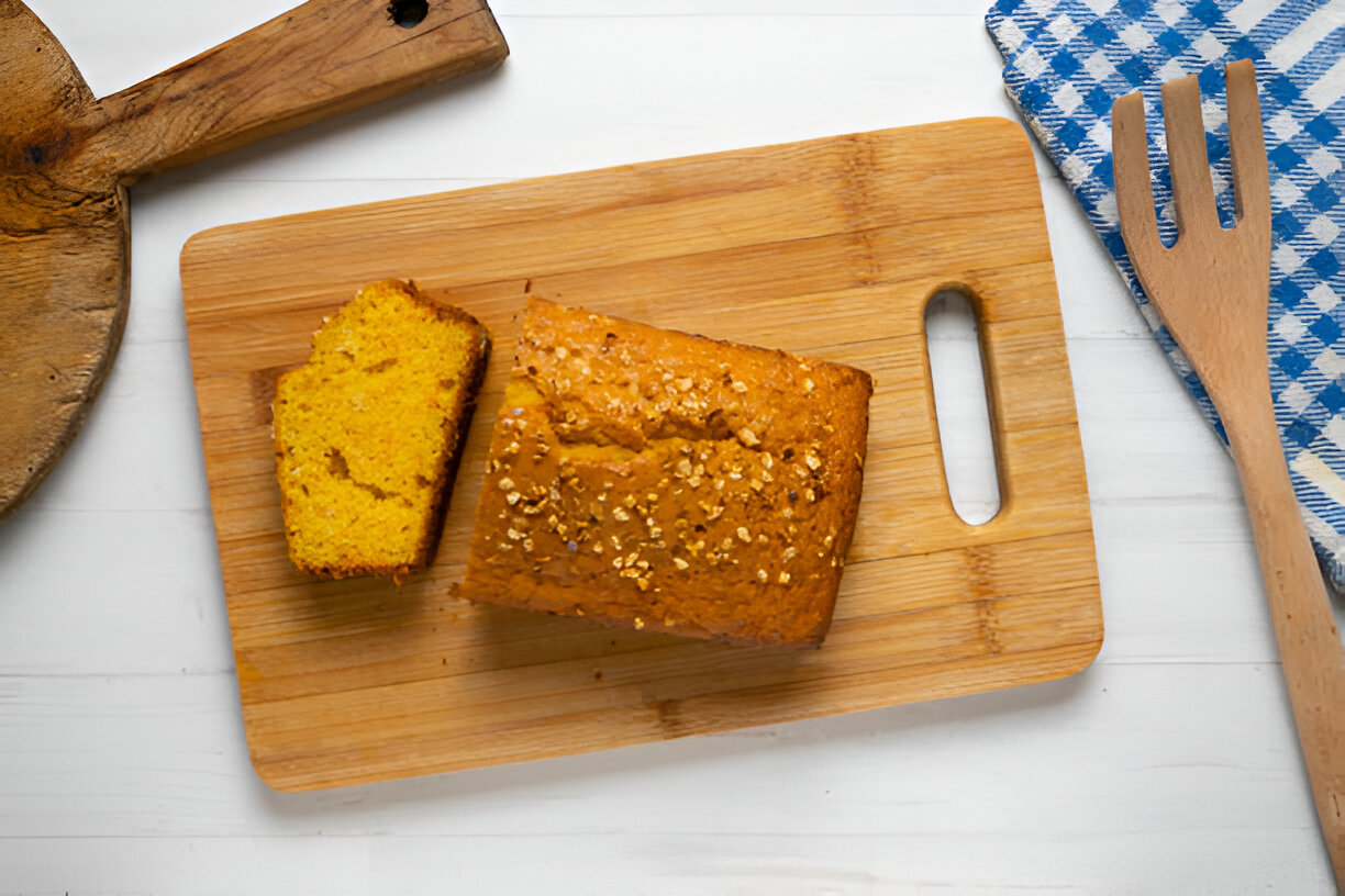 A freshly baked loaf of yellow bread with a slice cut off, resting on a wooden cutting board, accompanied by a blue and white checkered napkin and a wooden utensil.