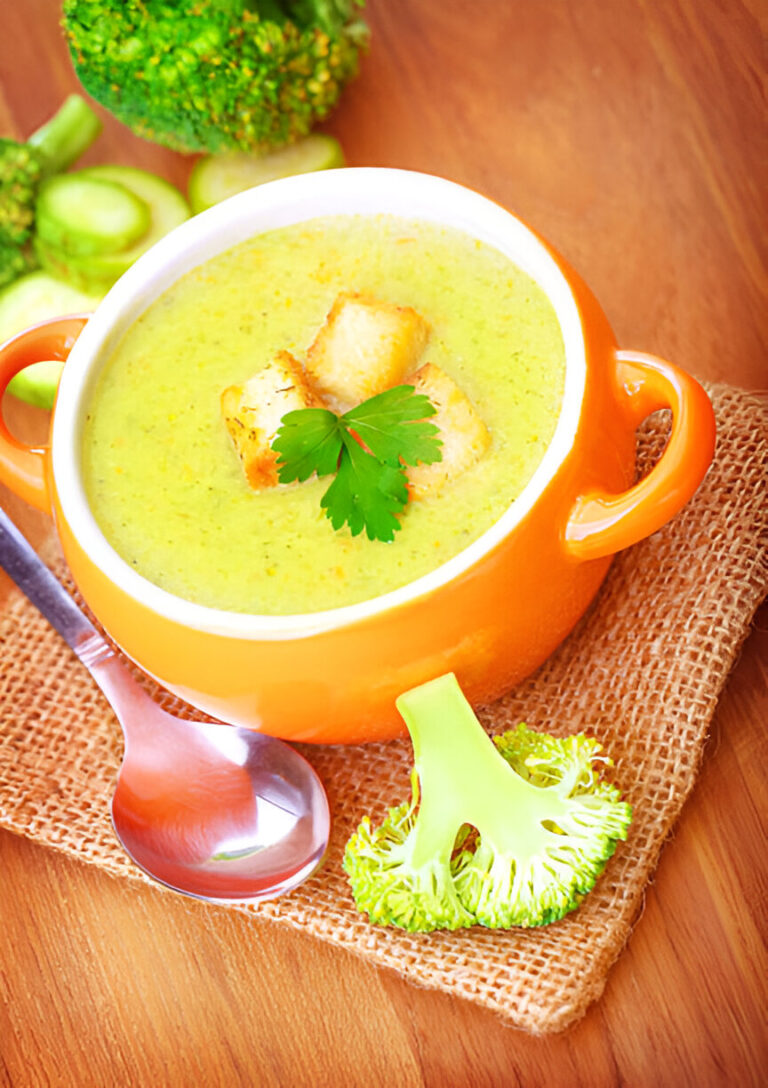 A bowl of creamy broccoli soup garnished with croutons and parsley, accompanied by fresh broccoli on a wooden surface.