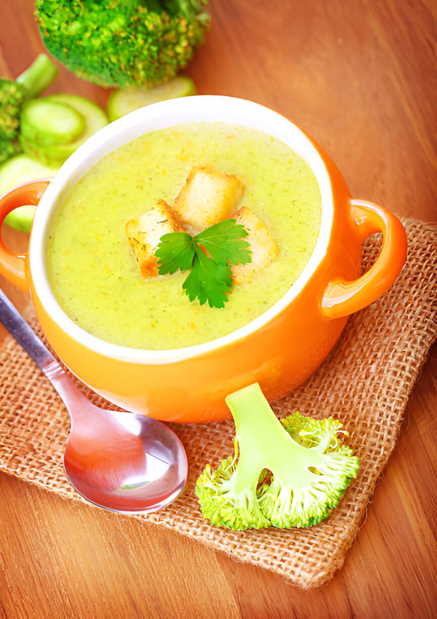 A bowl of creamy broccoli soup garnished with croutons and parsley, accompanied by fresh broccoli on a wooden surface.