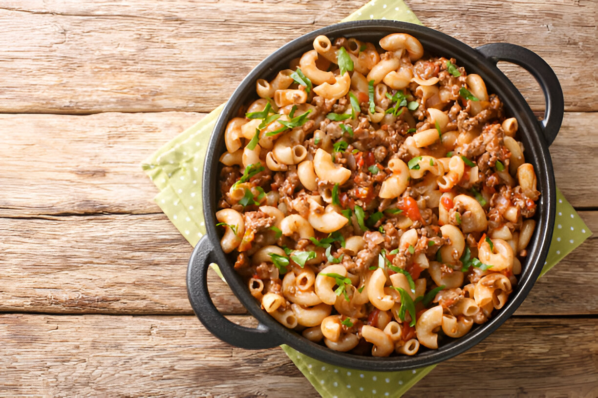 A pot of macaroni pasta with ground beef and green parsley on a wooden table.