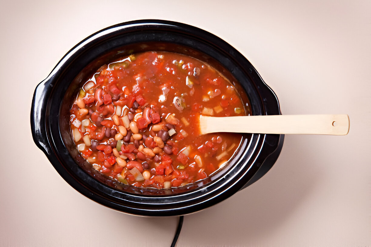 A black slow cooker filled with a colorful bean stew topped with vegetables and a wooden spoon.