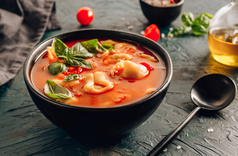 A bowl of tortellini soup garnished with fresh basil leaves, surrounded by cherry tomatoes and a bottle of olive oil.