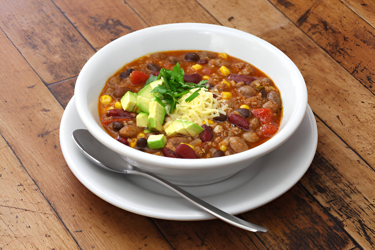 A bowl of hearty chili topped with diced avocado, shredded cheese, and chopped cilantro, served on a wooden table.