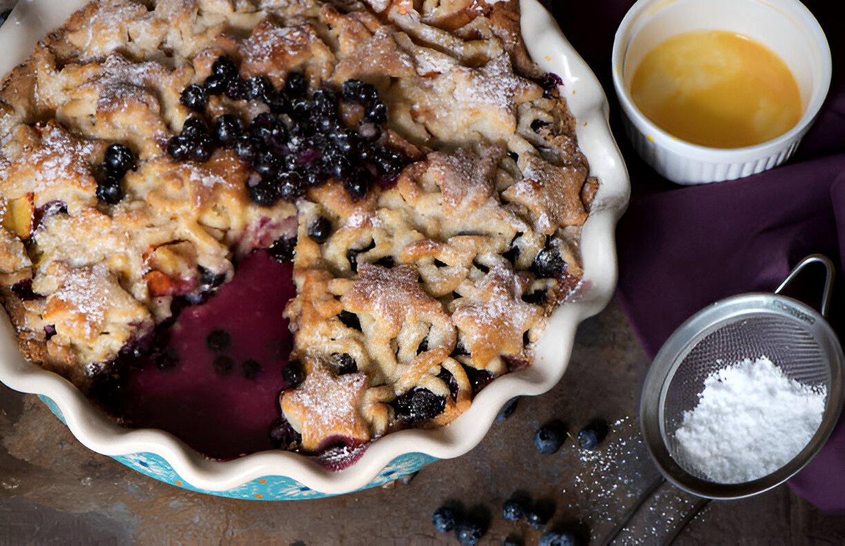 A freshly baked skillet pie filled with blueberries and a golden-brown crust, dusted with powdered sugar, next to a small bowl of melted butter and a sieve with powdered sugar.