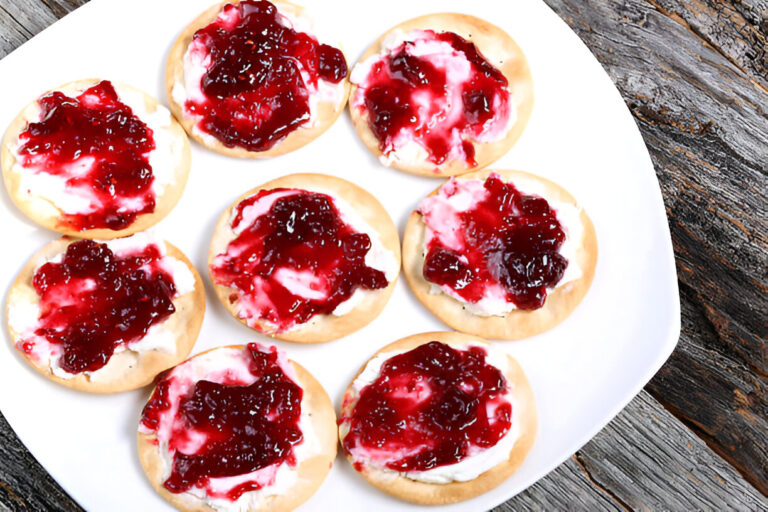 A white plate filled with round cookies topped with cream cheese and red fruit jam