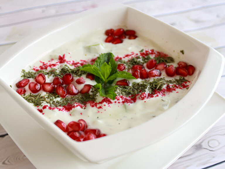 A creamy yogurt dish garnished with pomegranate seeds, mint leaves, and colorful spices, served in a white bowl.