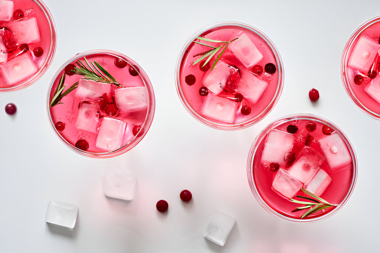 Five glasses of pink cocktails with ice cubes and garnished with cranberries and rosemary