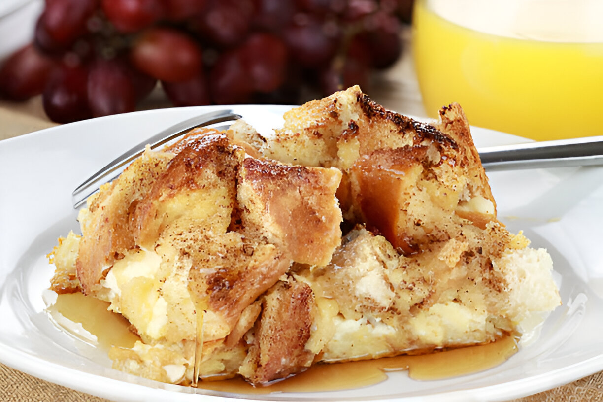 A slice of baked bread pudding topped with syrup on a white plate, with grapes and orange juice in the background.