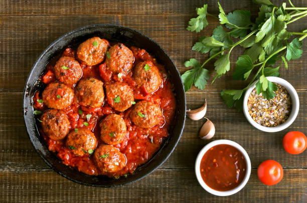 A skillet filled with meatballs in tomato sauce, garnished with parsley, alongside garlic, tomatoes, and spice bowls.