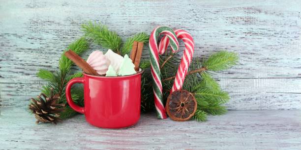 A festive red mug filled with colorful marshmallows, surrounded by candy canes and pine branches.