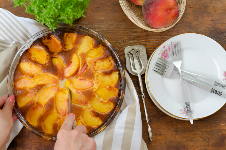 A freshly baked peach dessert surrounded by utensils and peaches on a wooden table.