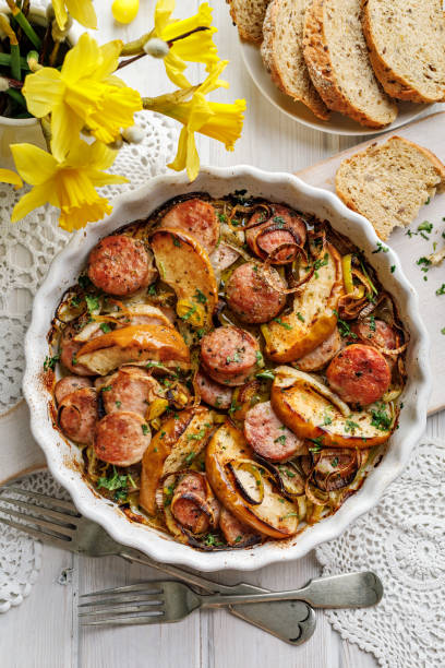 A delicious casserole dish with sliced sausages, potatoes, and onions garnished with herbs, served with slices of bread and yellow daffodils in the background.