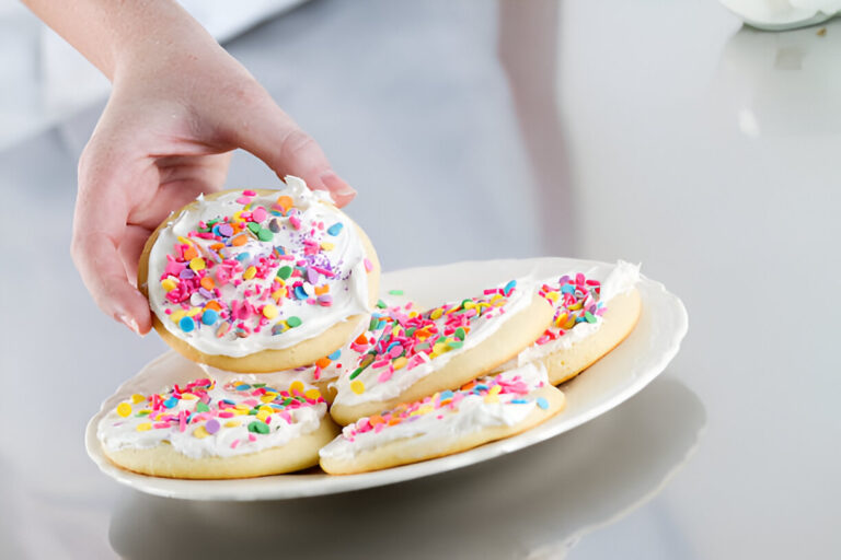 A hand picking up a frosted sugar cookie topped with colorful sprinkles from a plate.