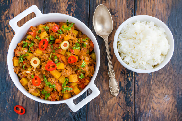 A bowl of colorful mixed vegetables and seasoned meat served alongside a bowl of white rice on a wooden table.