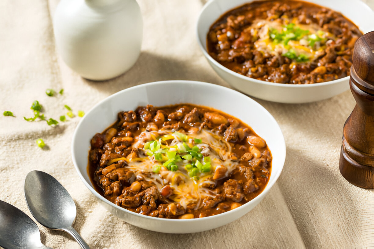 Two bowls of hearty chili topped with cheese and green onions, with silver spoons and a pepper mill in the background.