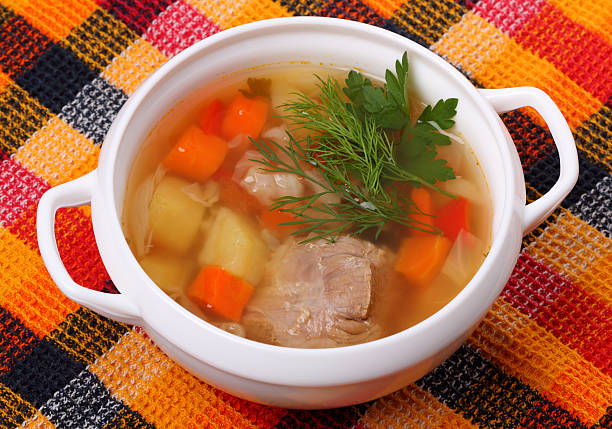 A white bowl of soup with vegetables and herbs on a colorful checked tablecloth.