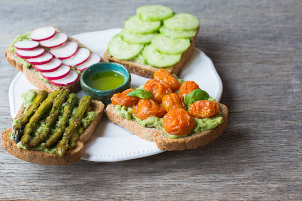 Four colorful slices of toast topped with various vegetables, including radishes, cucumbers, grilled asparagus, and cherry tomatoes, accompanied by a small dish of olive oil.