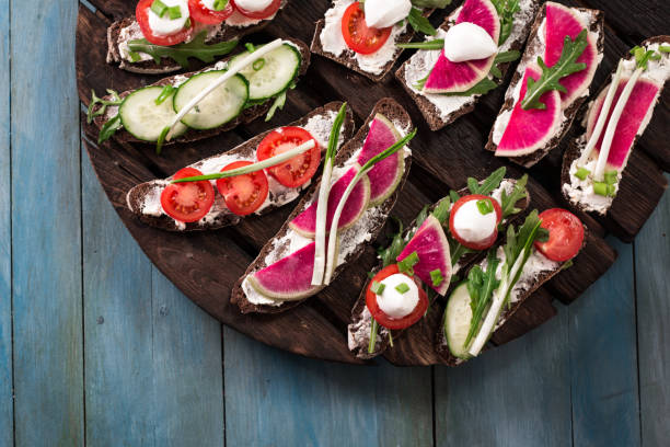 A wooden platter with open-faced sandwiches topped with various vegetables and herbs, including tomatoes, cucumbers, and radishes.