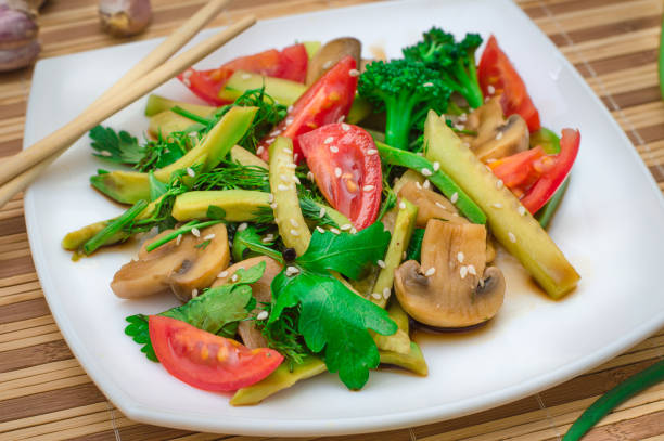 A colorful dish of sautéed vegetables including mushrooms, broccoli, and tomatoes, garnished with sesame seeds on a white plate.