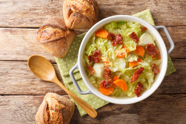 A bowl of cabbage soup with carrots and potatoes, accompanied by crusty bread rolls on a wooden table.