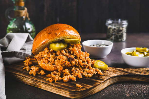 A close-up of a pulled meat sandwich with pickles on a wooden cutting board.