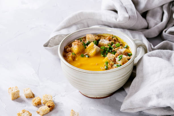 A bowl of creamy soup topped with croutons and green onions, set on a light background with some bread pieces scattered nearby.