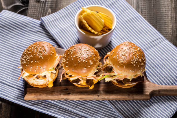 Three sesame seed topped sliders arranged on a wooden board with a side of potato chips in a cup