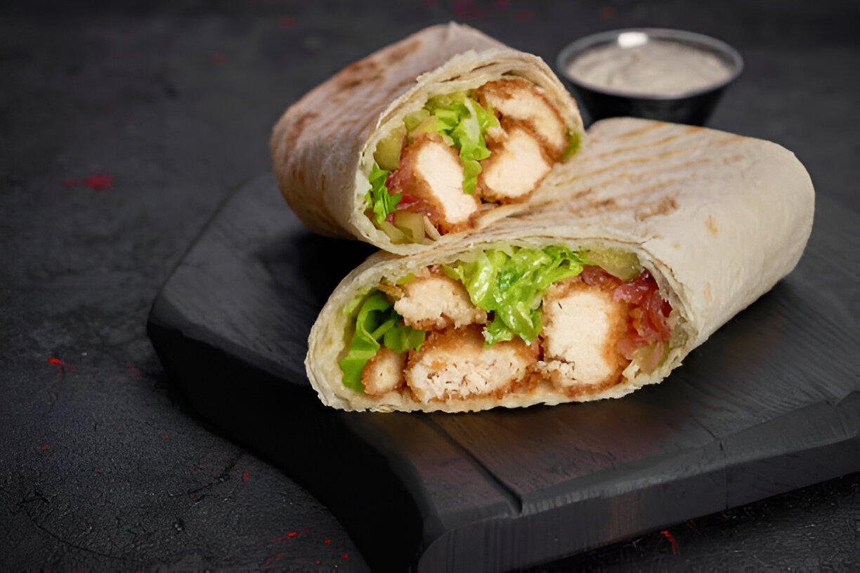 Close-up of a grilled chicken wrap filled with lettuce and sauces, served on a dark wooden board.