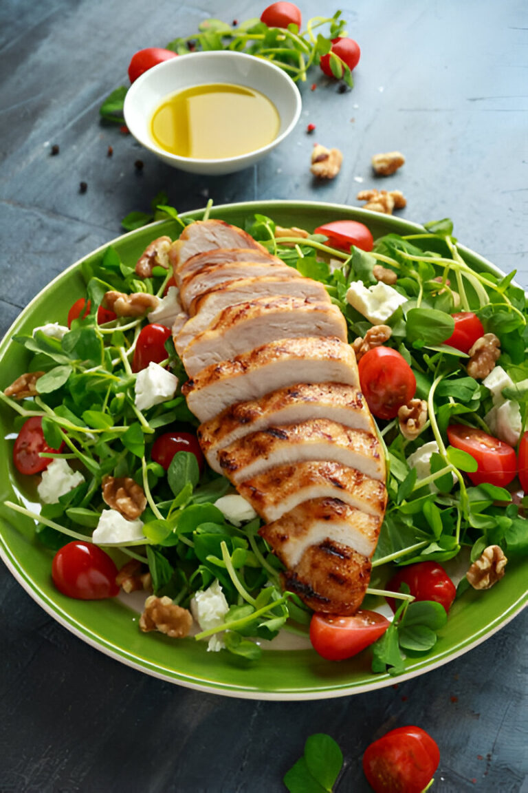 Grilled chicken breast served on a bed of mixed greens, cherry tomatoes, walnuts, and feta cheese, with olive oil in a bowl in the background.