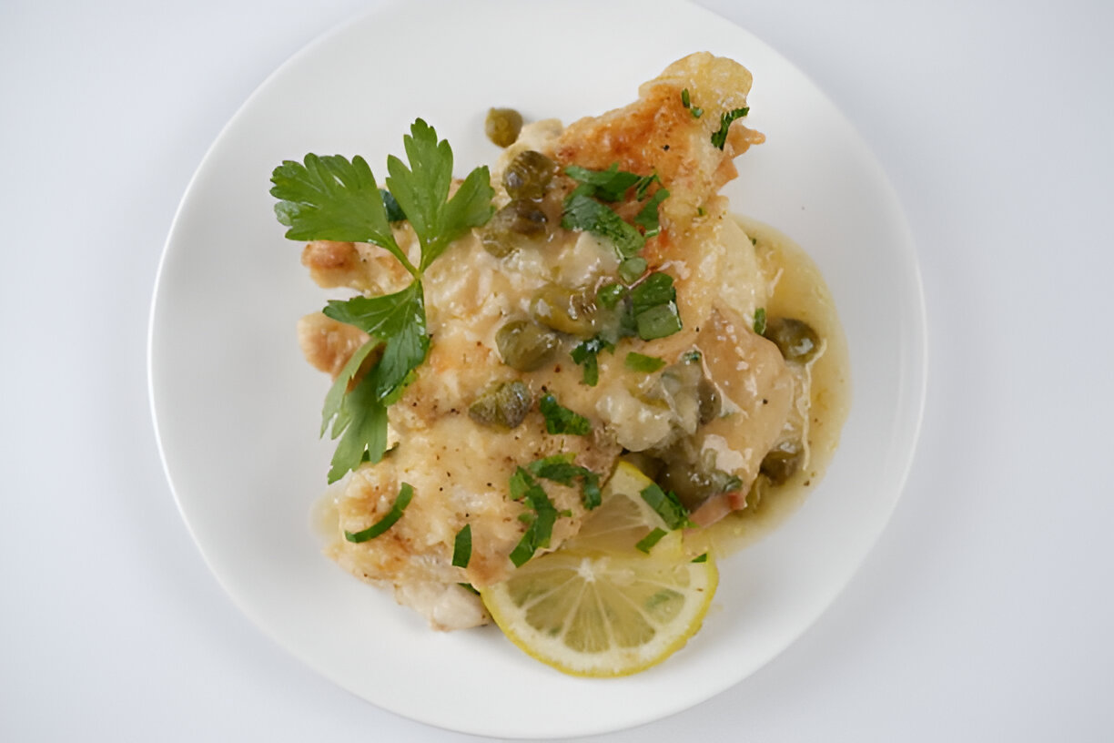 A plate of cooked fish topped with capers and herbs, garnished with lemon slices.