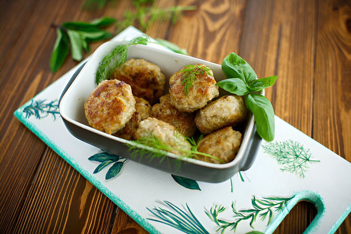 A serving of freshly made meatballs garnished with herbs in a bowl on a decorative cutting board.