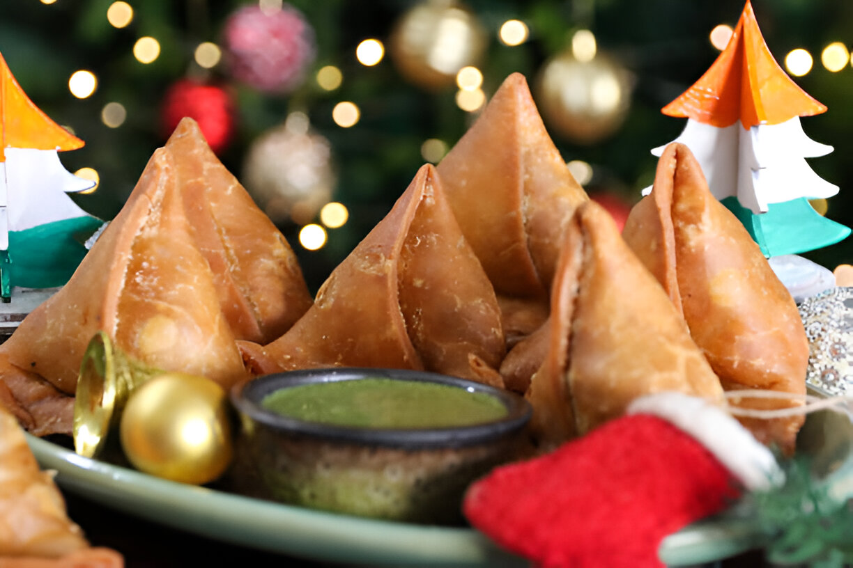 A plate of golden-brown samosas with a bowl of green chutney, decorated with festive ornaments and miniature Christmas trees in the background.