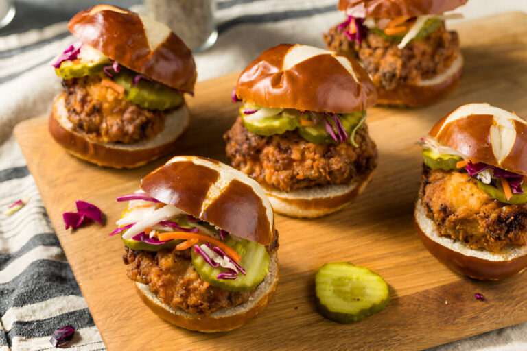 A wooden board displaying pretzel chicken sliders with pickles and colorful slaw.