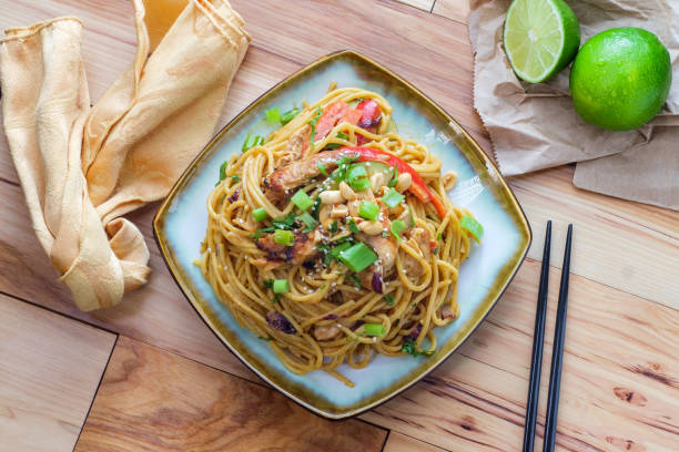 A plate of noodles with vegetables and chicken, garnished with green onions and served with a lime on the side.