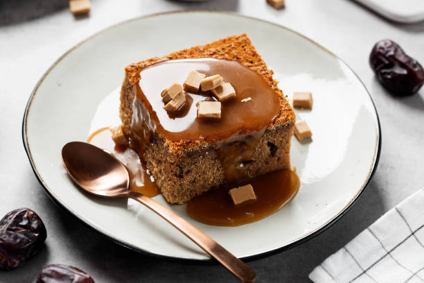 A piece of moist cake topped with caramel sauce and chocolate pieces on a white plate, next to a shiny spoon.