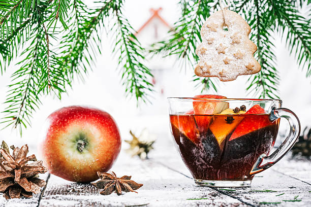 A glass of spiced fruit tea with a cookie shaped like a Christmas tree hanging from it, surrounded by apples and pine cones.