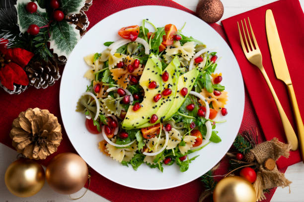 A colorful salad featuring farfalle pasta, avocado, pomegranate seeds, cherry tomatoes, and arugula, presented on a round white plate against a festive red background.