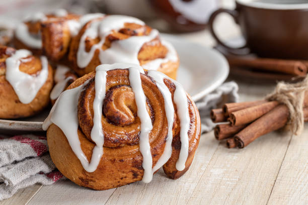 Freshly baked cinnamon rolls drizzled with icing on a plate, with cinnamon sticks and a cup in the background.