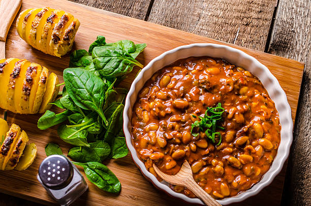 A bowl of baked beans topped with chopped green onions, served with spinach and baked potatoes on a wooden surface.