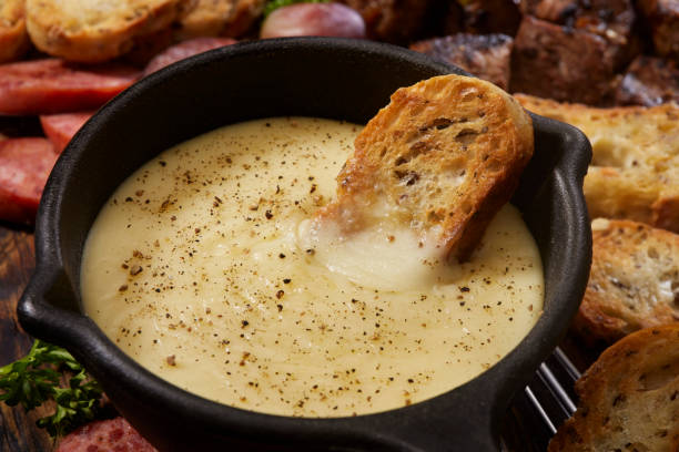 A serving of creamy cheese fondue in a black pot with a bread slice dipped into it, surrounded by assorted meats and toasted bread.