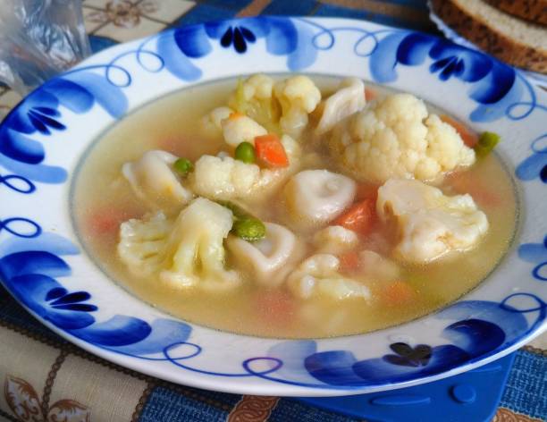 A bowl of vegetable soup featuring cauliflower, carrots, and green peas in a clear broth on a decorative plate.