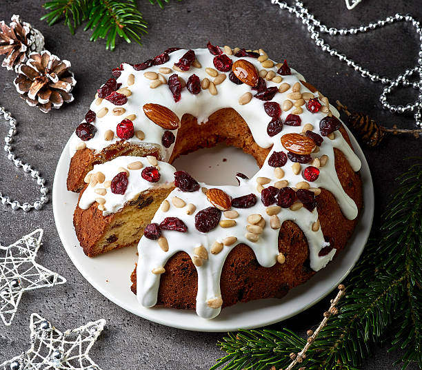 A festive bundt cake decorated with white icing, almonds, cranberries, and pine nuts on a white plate, surrounded by holiday decorations.